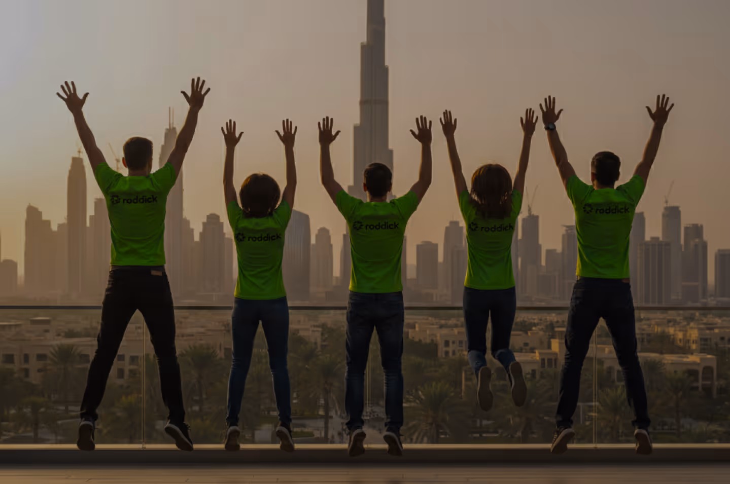 Five people wearing green 'roddick' shirts jumping with hands raised on a balcony overlooking a city skyline at sunset.