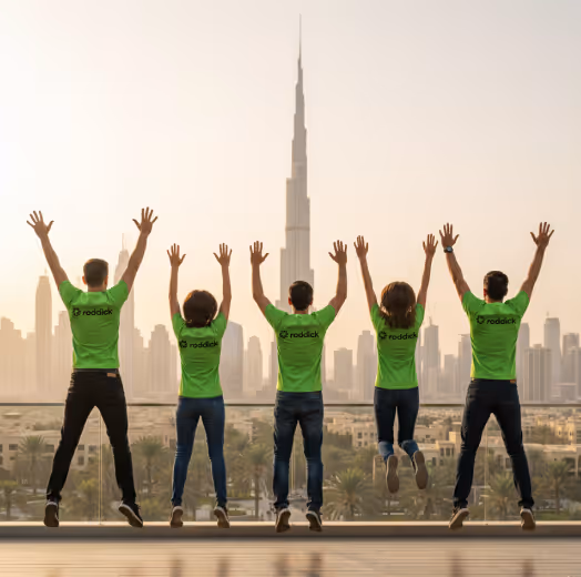 Five people wearing green shirts jumping with raised hands in front of Dubai skyline with Burj Khalifa.